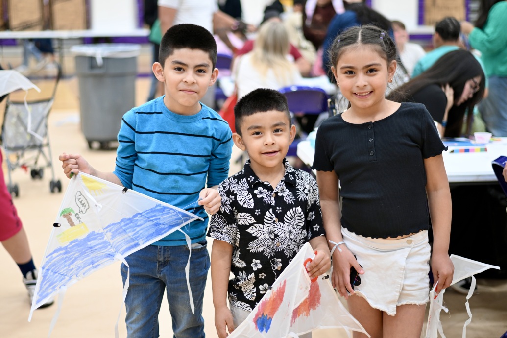 three students with kites that they drew on, smiling in a gym